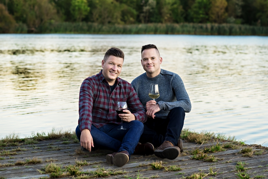 An engaged couple sit on a dock next to a lake in FDR Park holding glasses of wine.